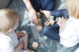 Two young children are sitting on a textured blue rug, playing with colorful letter blocks. An adult's hand is arranging some of the blocks, engaging with the children. The children appear to be focused on the activity, and the scene is well-lit with natural light.
