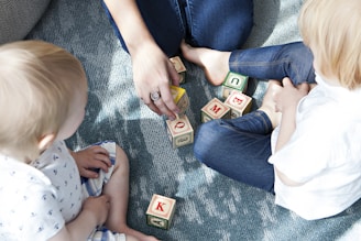 Two young children are sitting on a textured blue rug, playing with colorful letter blocks. An adult's hand is arranging some of the blocks, engaging with the children. The children appear to be focused on the activity, and the scene is well-lit with natural light.