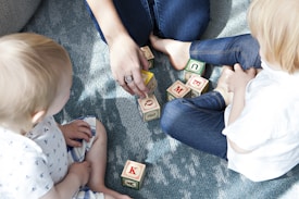 Two young children are sitting on a textured blue rug, playing with colorful letter blocks. An adult's hand is arranging some of the blocks, engaging with the children. The children appear to be focused on the activity, and the scene is well-lit with natural light.