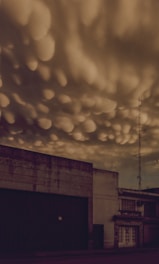 Dark and dramatic skies with unusual cloud formations hover over an industrial building. The clouds exhibit a bulbous texture, adding a sense of foreboding to the scene. A tall antenna tower stands beside the building, silhouetted against the ominous sky.