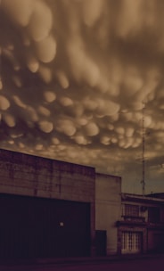 Dark and dramatic skies with unusual cloud formations hover over an industrial building. The clouds exhibit a bulbous texture, adding a sense of foreboding to the scene. A tall antenna tower stands beside the building, silhouetted against the ominous sky.