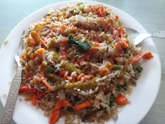 A plate of fried rice with mixed vegetables and sauces, garnished with strips of carrots and green beans. The dish is served on a white plate with a fork and spoon on either side.