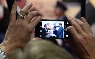 A close-up shot of hands holding a smartphone displaying a photograph of a group of people. The image on the phone shows individuals dressed in formal attire indoors, possibly at an event or gathering. The focus on the phone suggests capturing a moment of significance.