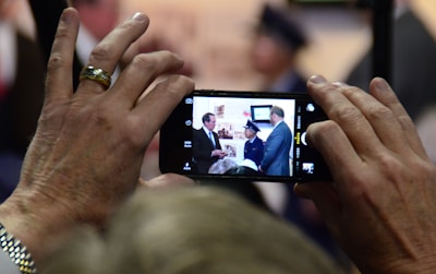 A close-up shot of hands holding a smartphone displaying a photograph of a group of people. The image on the phone shows individuals dressed in formal attire indoors, possibly at an event or gathering. The focus on the phone suggests capturing a moment of significance.