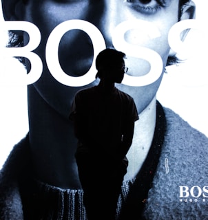 A silhouette of a person stands in front of a large advertisement featuring part of a black-and-white face with the word 'BOSS' prominently displayed. The figure is dark against the brightly lit background.