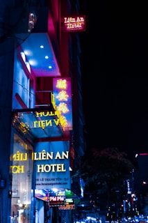 Neon signs of a hotel appear prominently against the dark night sky. The signs are vibrant with yellow and pink hues, reflecting onto the glass facade. The street appears lively with additional neon lights in the background.