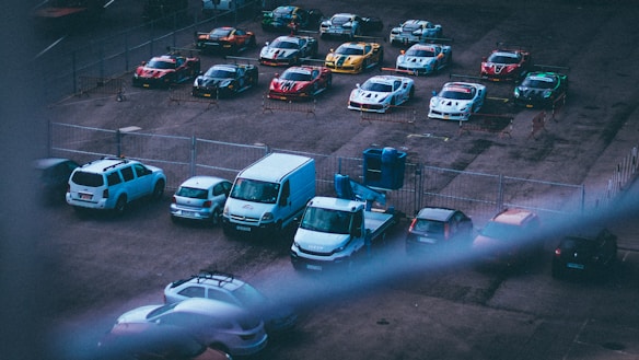 A fenced parking lot contains a row of sports cars, each marked with racing numbers and advertisements. In front of the sports cars, several ordinary vehicles are parked, including a few vans and sedans. The lighting suggests an early morning or late afternoon setting, with shadows indicating a low sun angle.