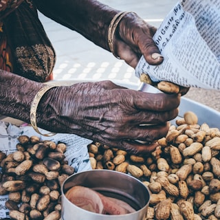 A pair of elderly hands are wrapping peanuts in a newspaper cone. The hands are adorned with gold bangles, showing signs of age and experience. There is a pile of peanuts on a table, and a metal container with currency notes next to it.