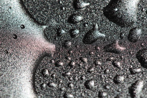 Shimmering texture with sparkling droplets scattered across a metallic surface. The surface reflects iridescent colors, creating a visually dynamic scene.