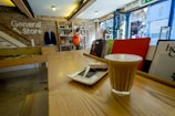 A cozy store interior with rustic brick walls and wooden counters. A latte and a chocolate square are served on a small tray in the foreground. A person with an orange backpack browses in the background. Shelves display various books and items, and sunlight filters through the open glass door.