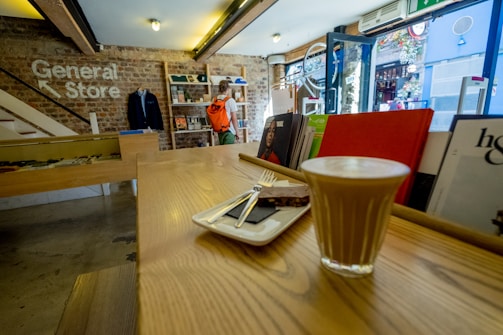 A cozy store interior with rustic brick walls and wooden counters. A latte and a chocolate square are served on a small tray in the foreground. A person with an orange backpack browses in the background. Shelves display various books and items, and sunlight filters through the open glass door.