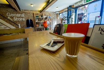 A cozy store interior with rustic brick walls and wooden counters. A latte and a chocolate square are served on a small tray in the foreground. A person with an orange backpack browses in the background. Shelves display various books and items, and sunlight filters through the open glass door.