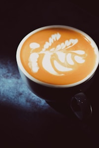 A cup of coffee with intricate latte art on its surface, featuring a delicate leaf pattern. The cup is placed on a dark surface with a soft light illuminating the design, creating a warm and inviting atmosphere.