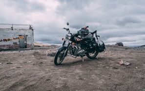 A motorcycle is parked on a barren, rocky terrain under an overcast sky. The motorcycle is equipped with bags and gear, suggesting a journey or expedition. In the background, there is a dilapidated structure with colorful prayer flags attached to it, adding a touch of color to the otherwise muted landscape. A vehicle is visible in the distance, possibly on a dirt road.