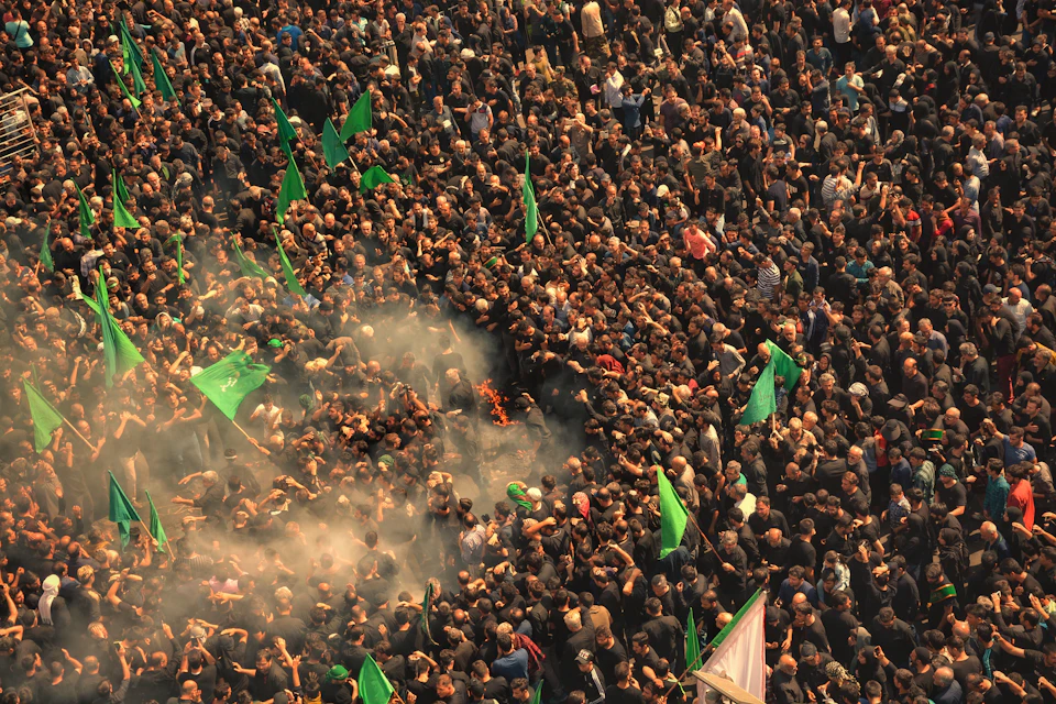 The image depicts a large crowd of people gathered together, with many individuals holding green flags and banners. The scene is set against an urban backdrop, suggesting that it may be taking place in a city or town center. The perspective of the photo is from above, providing a bird's eye view of the event.