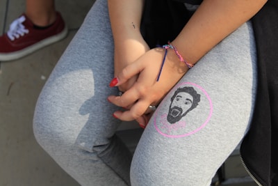 A person wearing grey leggings with a printed face on one leg and holding their hands in front, showing red nail polish. A colorful friendship bracelet is on their wrist, and they are seated with a visible red sneaker in the background.
