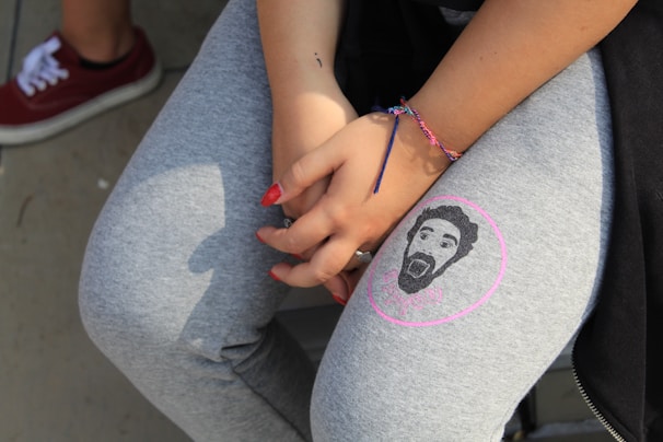 A person wearing grey leggings with a printed face on one leg and holding their hands in front, showing red nail polish. A colorful friendship bracelet is on their wrist, and they are seated with a visible red sneaker in the background.