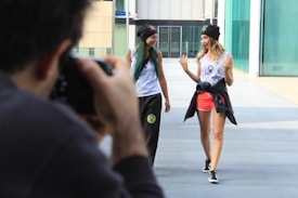 Two women walk side by side on a modern walkway, laughing and engaging in conversation. Both are wearing casual athletic clothing, including beanies and tank tops, and one woman has a jacket tied around her waist. In the foreground, a man captures the moment with a camera, slightly blurred out to emphasize focus on the women. The background features contemporary architecture with large glass panels.