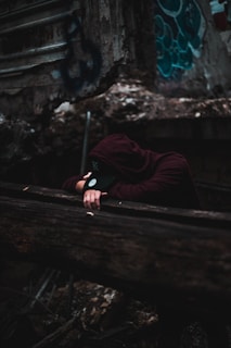 A person wearing a dark hoodie and cap is leaning over a wooden railing, seemingly in a state of exhaustion or contemplation. The background features old, rough, and graffiti-marked walls, suggesting an urban or abandoned setting.