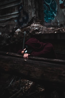 A person wearing a dark hoodie and cap is leaning over a wooden railing, seemingly in a state of exhaustion or contemplation. The background features old, rough, and graffiti-marked walls, suggesting an urban or abandoned setting.