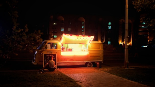 A brightly lit food truck with a yellow glow sits on a dark street at night. A person is sitting on the ground nearby, possibly enjoying their purchase. The truck is equipped with a menu board and string lights, creating a warm and inviting ambiance against the dark background. Buildings with illuminated windows can be seen in the distance.