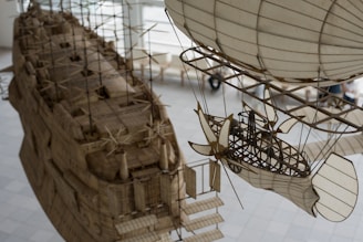 An intricate and detailed model of an airship and a ship are suspended indoors. The design features various structural elements such as propellers, support beams, and sails, all crafted with precision to resemble retro-futuristic or steampunk-style vehicles.