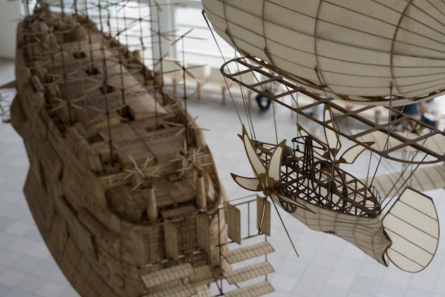 An intricate and detailed model of an airship and a ship are suspended indoors. The design features various structural elements such as propellers, support beams, and sails, all crafted with precision to resemble retro-futuristic or steampunk-style vehicles.