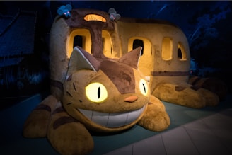 A large, plush sculpture depicting a whimsical cat-bus hybrid from an animated film. The creature has a big, friendly grin, bright glowing eyes, and is colored in shades of brown and beige with stripes. There are mouse-like creatures on its back, and windows along its sides indicate that it functions as a bus.