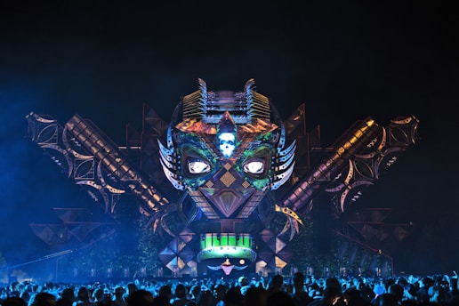 An intricately designed stage at a festival features a large, illuminated owl face with elaborate patterns and mechanical wings extending outward. The stage is decorated with vibrant lights and geometric shapes, creating a mystical and futuristic atmosphere. A large crowd of people is gathered in front, silhouetted against the colorful lights.