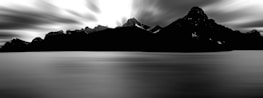 A serene, long-exposure photograph of a mountain range silhouetted against a dramatic sky. The water in the foreground is smooth and reflective, creating a tranquil and ethereal atmosphere. The high contrast between the dark mountains and the bright sky adds depth and intensity to the scene.