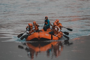 A group of people wearing helmets and life jackets paddle together on an orange inflatable raft in a calm body of water.