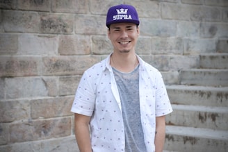 A young man is standing in front of a stone wall and steps. He is wearing a purple cap with the word SUPRA and a crown logo, a white patterned short-sleeve shirt, and a gray t-shirt underneath. He is smiling and looking directly at the camera.