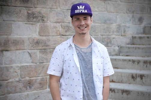 A young man is standing in front of a stone wall and steps. He is wearing a purple cap with the word SUPRA and a crown logo, a white patterned short-sleeve shirt, and a gray t-shirt underneath. He is smiling and looking directly at the camera.