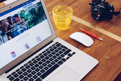 A MacBook Air displaying a webpage about Facebook ads is on a wooden desk. Next to the laptop is a white mouse, a red pencil, a glass filled with yellow plastic stirrers, and a black camera.