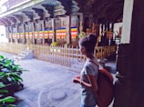 A person stands in a historical or cultural site with intricate architectural details and colorful flags. The area is fenced with gold-colored railings and there are religious or ceremonial objects, alongside lush greenery.