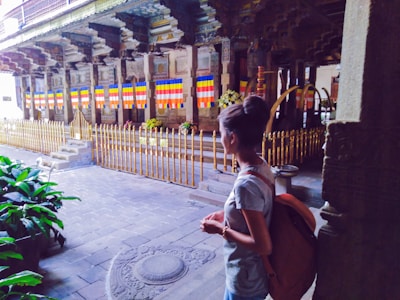 A person stands in a historical or cultural site with intricate architectural details and colorful flags. The area is fenced with gold-colored railings and there are religious or ceremonial objects, alongside lush greenery.
