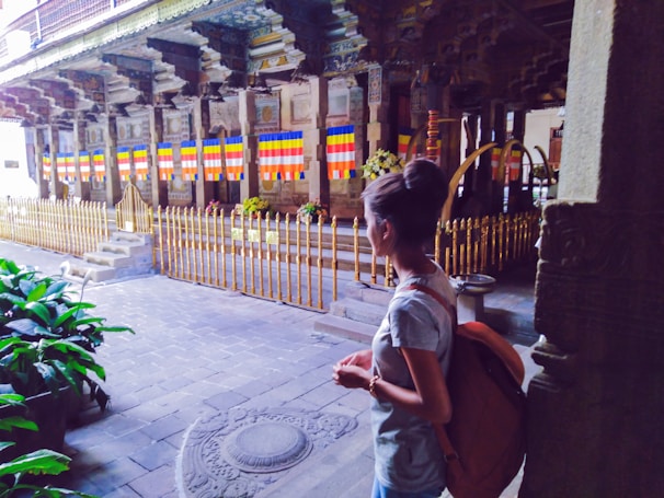 A person stands in a historical or cultural site with intricate architectural details and colorful flags. The area is fenced with gold-colored railings and there are religious or ceremonial objects, alongside lush greenery.