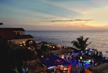 An outdoor seaside venue with a view of the ocean at sunset. There are several people gathered around tables and standing in groups, and the area is illuminated with ambient lights and colorful spotlights. A palm tree adds a tropical feel to the setting. The sky is painted with soft hues of orange, pink, and blue as the sun sets over the horizon.