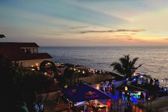 An outdoor seaside venue with a view of the ocean at sunset. There are several people gathered around tables and standing in groups, and the area is illuminated with ambient lights and colorful spotlights. A palm tree adds a tropical feel to the setting. The sky is painted with soft hues of orange, pink, and blue as the sun sets over the horizon.