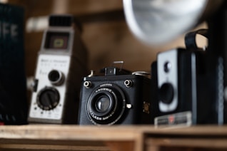 A collection of vintage cameras is displayed on a wooden shelf. The focus is on the central camera, an old box camera with a prominent lens and textured exterior. Other cameras are visible in the background, showcasing different designs and styles. The lighting is soft, emphasizing the nostalgic quality of the scene.