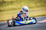 A child is driving a go-kart on a racetrack. The driver wears a white racing helmet and a blue racing suit. The go-kart displays the number 15 on the front and is primarily blue, yellow, and white in color. The background features a grassy area beside the track.