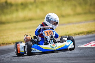 A child is driving a go-kart on a racetrack. The driver wears a white racing helmet and a blue racing suit. The go-kart displays the number 15 on the front and is primarily blue, yellow, and white in color. The background features a grassy area beside the track.