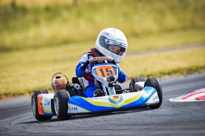 A child is driving a go-kart on a racetrack. The driver wears a white racing helmet and a blue racing suit. The go-kart displays the number 15 on the front and is primarily blue, yellow, and white in color. The background features a grassy area beside the track.