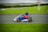 A person wearing a helmet and racing gear is driving a go-kart on an outdoor track. The racer is moving at high speed, captured in motion with a blurred background. The track is surrounded by green grass, and there are distant hills and cloudy skies in the background.