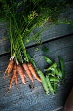 A bunch of freshly harvested carrots and snap peas are laid out on a rustic wooden surface. The carrots have vibrant green tops, and the peas are enclosed in their pods.