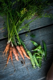 A bunch of freshly harvested carrots and snap peas are laid out on a rustic wooden surface. The carrots have vibrant green tops, and the peas are enclosed in their pods.