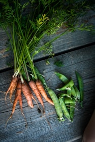 A bunch of freshly harvested carrots and snap peas are laid out on a rustic wooden surface. The carrots have vibrant green tops, and the peas are enclosed in their pods.