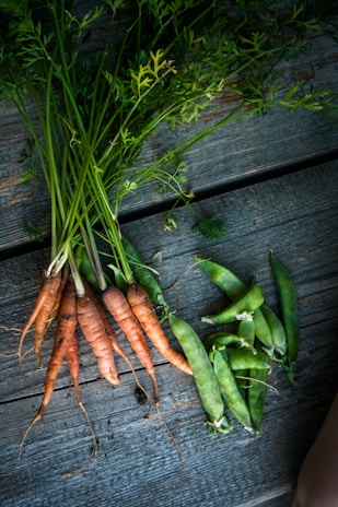 A bunch of freshly harvested carrots and snap peas are laid out on a rustic wooden surface. The carrots have vibrant green tops, and the peas are enclosed in their pods.