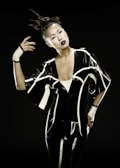 A person stands against a dark background wearing a futuristic, glossy black outfit with white geometric lines. The hair is styled in an avant-garde topknot, and the makeup is bold with dark lipstick.