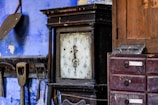 A vintage, ornate clock with roman numerals is set against a rustic backdrop. On one side, wooden tools hang against a worn blue wall, while on the other, a wooden cabinet with labeled drawers sits. The overall setting suggests an old workshop or a quaint antique space.